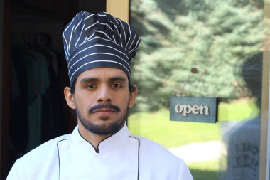 Yakışıklı Latin aşçının portresi. Üniformalı ve şapkalı. Restoran kapısının önünde.