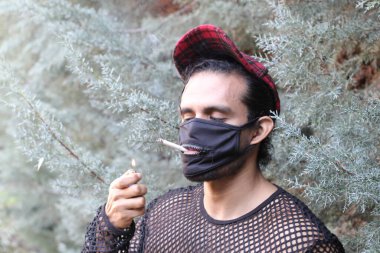 Açık havada sigara içen, net tişörtlü ve maskeli genç adamın yakın plan portresi.