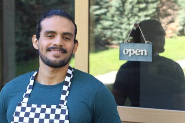 Kapının önünde açık hesap ile damalı önlüklü yakışıklı Latin küçük işletme sahibinin portresi.