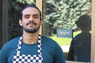 Kapının önünde açık hesap ile damalı önlüklü yakışıklı Latin küçük işletme sahibinin portresi.