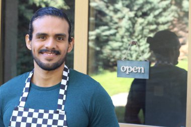 Kapının önünde açık hesap ile damalı önlüklü yakışıklı Latin küçük işletme sahibinin portresi.