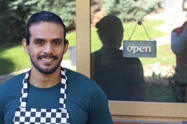 Kapının önünde açık hesap ile damalı önlüklü yakışıklı Latin küçük işletme sahibinin portresi.