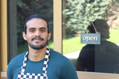 Kapının önünde açık hesap ile damalı önlüklü yakışıklı Latin küçük işletme sahibinin portresi.