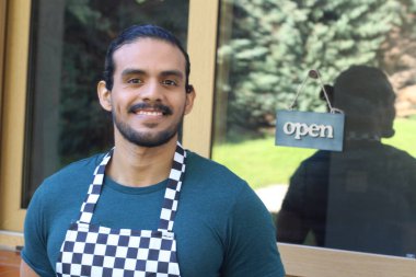 Kapının önünde açık hesap ile damalı önlüklü yakışıklı Latin küçük işletme sahibinin portresi.