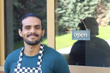 Kapının önünde açık hesap ile damalı önlüklü yakışıklı Latin küçük işletme sahibinin portresi.