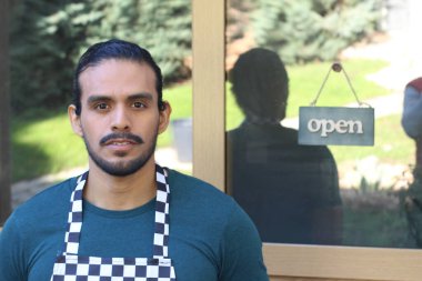 Kapının önünde açık hesap ile damalı önlüklü yakışıklı Latin küçük işletme sahibinin portresi.