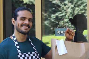 Kapının önünde kağıt torba ve açık hesap ile yakışıklı Latin üniformalı küçük işletme sahibi portresi