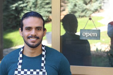 Kapının önünde açık hesap ile damalı önlüklü yakışıklı Latin küçük işletme sahibinin portresi.