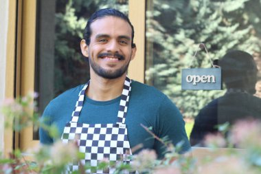 Kapının önünde açık hesap ile damalı önlüklü yakışıklı Latin küçük işletme sahibinin portresi.