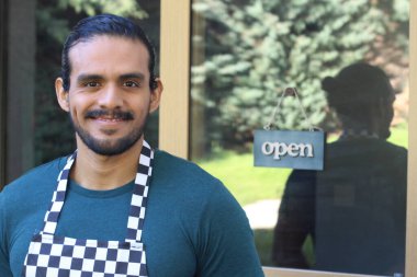 Kapının önünde açık hesap ile damalı önlüklü yakışıklı Latin küçük işletme sahibinin portresi.