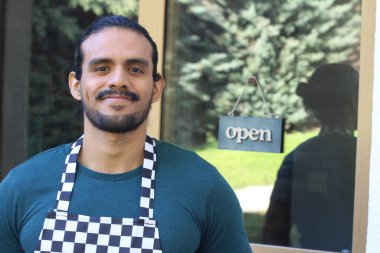 Kapının önünde açık hesap ile damalı önlüklü yakışıklı Latin küçük işletme sahibinin portresi.