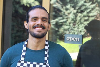 Kapının önünde açık hesap ile damalı önlüklü yakışıklı Latin küçük işletme sahibinin portresi.