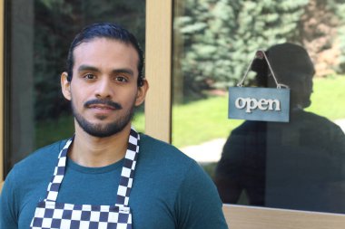 Kapının önünde açık hesap ile damalı önlüklü yakışıklı Latin küçük işletme sahibinin portresi.