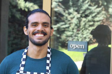 Kapının önünde açık hesap ile damalı önlüklü yakışıklı Latin küçük işletme sahibinin portresi.