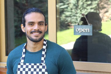 Kapının önünde açık hesap ile damalı önlüklü yakışıklı Latin küçük işletme sahibinin portresi.