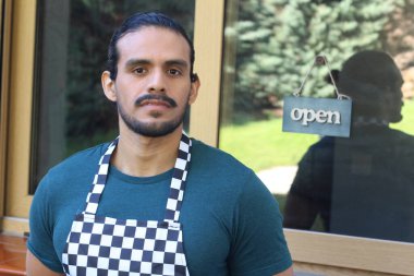 Kapının önünde açık hesap ile damalı önlüklü yakışıklı Latin küçük işletme sahibinin portresi.