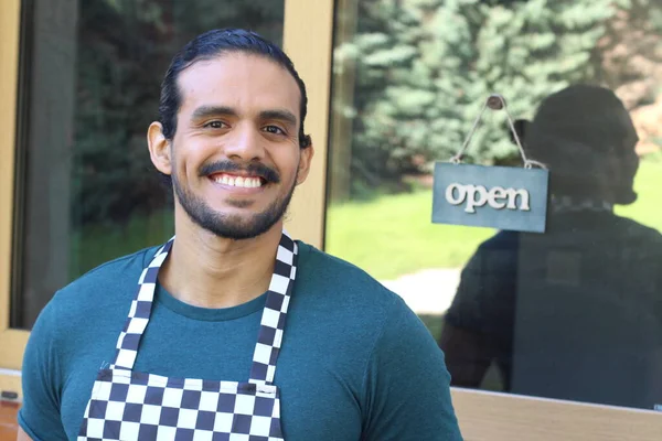 Kapının önünde açık hesap ile damalı önlüklü yakışıklı Latin küçük işletme sahibinin portresi.