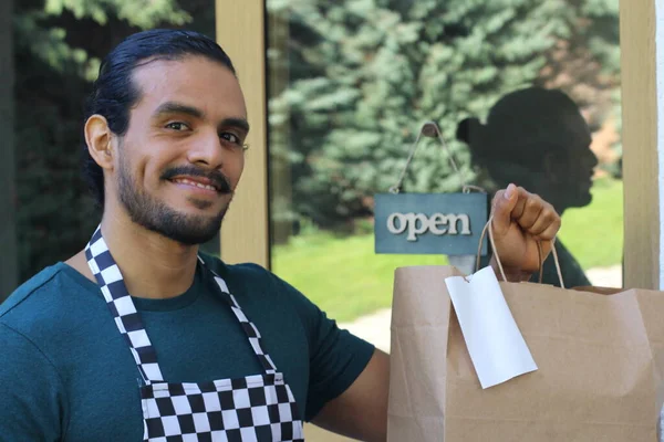 Kapının önünde kağıt torba ve açık hesap ile yakışıklı Latin üniformalı küçük işletme sahibi portresi