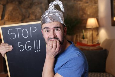 Alüminyum folyo şapkalı yakışıklı genç adam elinde karatahtayla evde 5G yazıtlar tutuyor.