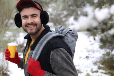 Kış ormanında sırt çantası ve kağıt kahve taşıyan yakışıklı genç bir adam.