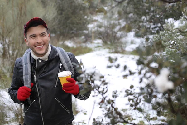 handsome young man in leather jacket with backpack and paper coffee cup in winter forest