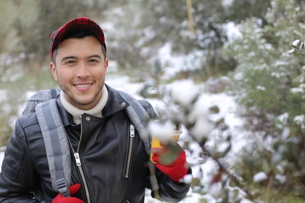 handsome young man in leather jacket with backpack and paper coffee cup in winter forest