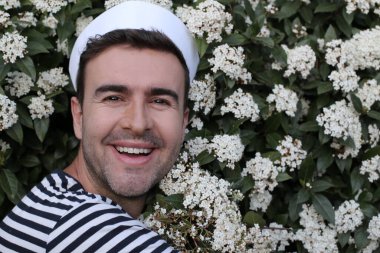 Beyaz çiçeklerle dolu çalıların önündeki denizci şapkalı yakışıklı genç adamın yakın plan portresi.