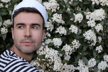 Beyaz çiçeklerle dolu çalıların önündeki denizci şapkalı yakışıklı genç adamın yakın plan portresi.