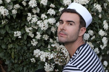 Beyaz çiçeklerle dolu çalıların önündeki denizci şapkalı yakışıklı genç adamın yakın plan portresi.