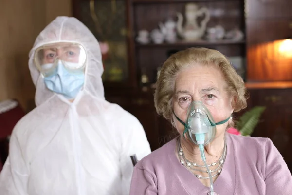 portrait of two mature woman in oxygen mask with doctor in protective costume at home, covid-19 concept