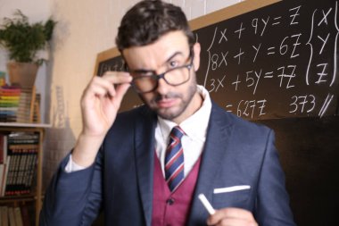 Sınıfta tahtaya tebeşirle tebeşir tutan takım elbiseli yakışıklı bir öğretmenin yakın plan portresi.