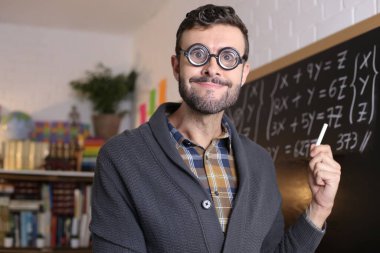 close-up portrait of handsome young teacher with funny eyeglasses holding chalk in front of blackboard in classroom
