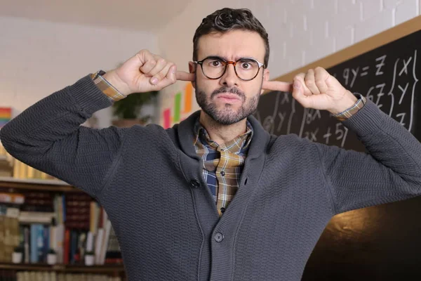close-up portrait of handsome young teacher covering ears with fingers in front of blackboard in classroom