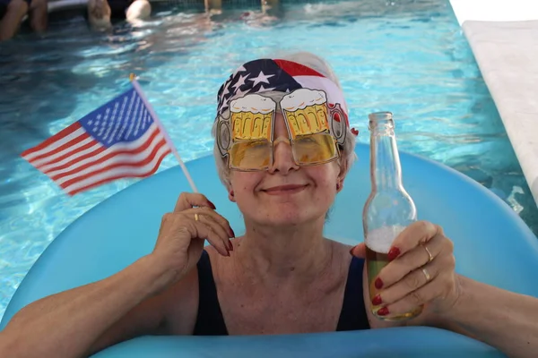 portrait of mature woman in swimsuit with usa flag, band and beer in pool on sunny summer day