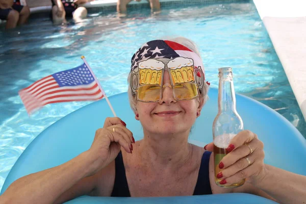 portrait of mature woman in swimsuit with usa flag, band and beer in pool on sunny summer day