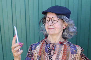 Yeşil duvarın önünde akıllı telefon kullanan şık giyinmiş güzel olgun bir kadının yakın plan portresi.