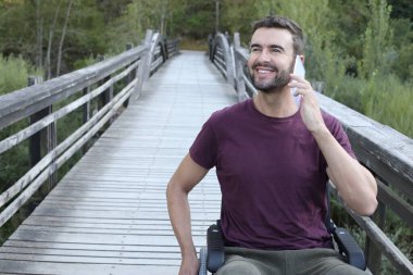 Parktaki ahşap köprüde telefonla konuşan tekerlekli sandalyedeki yakışıklı genç adamın portresi.