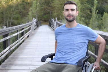 Parktaki ahşap köprüde tekerlekli sandalyedeki yakışıklı genç adamın portresi.