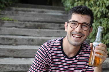 Yakından çekilmiş bir portre, sıradan kıyafetler giymiş yakışıklı bir adam merdivenlerde oturuyor ve elinde bira şişesi var.