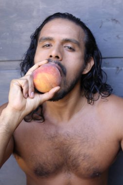 Yakışıklı, genç, melez bir adamın beton duvarın önünde şeftali yerken çekilmiş portresi.