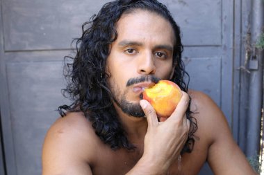 Yakışıklı, genç, melez bir adamın beton duvarın önünde şeftali yerken çekilmiş portresi.