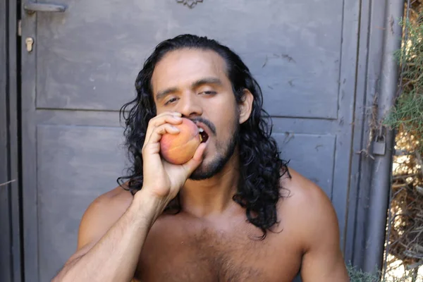 年轻帅气的混血男子赤身裸体在水泥墙前吃成熟的桃子的特写 — 图库照片
