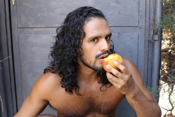 コンクリートの壁の前で熟れた桃を食べるハンサムな若い混合人種の恥知らずの男のクローズアップ肖像画 — ストック写真