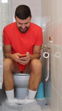 Genç adam akıllı telefonuyla tuvalette oturuyor..