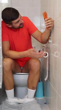 Genç adam akıllı telefonuyla tuvalette oturuyor..