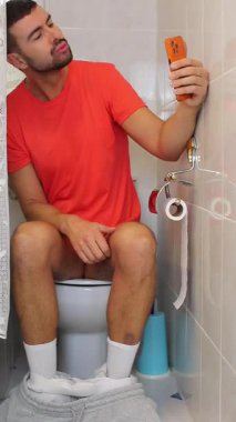 Genç adam akıllı telefonuyla tuvalette oturuyor..