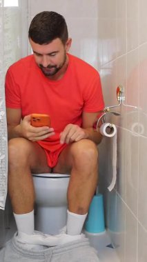 Genç adam akıllı telefonuyla tuvalette oturuyor..