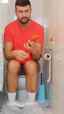 Genç adam akıllı telefonuyla tuvalette oturuyor..