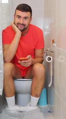 Genç adam akıllı telefonuyla tuvalette oturuyor..