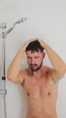 Genç erkek yetişkin modern duşta akan suyun altında saç yıkıyor.